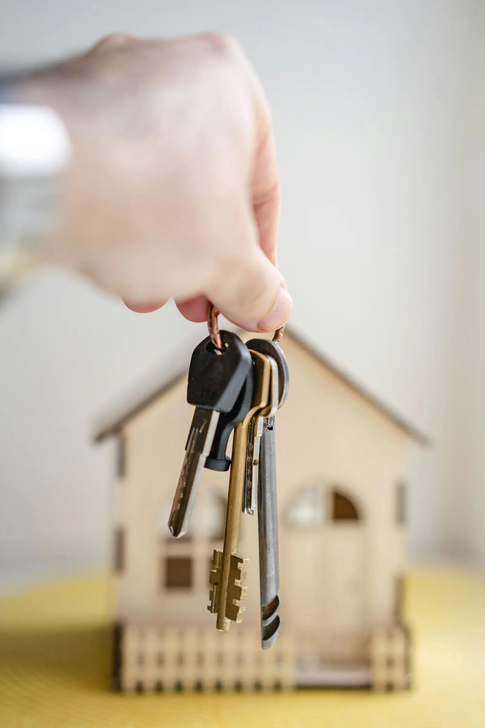 Close up of a hand holding keys with a miniature wooden house in the background symbolizing real estate investment