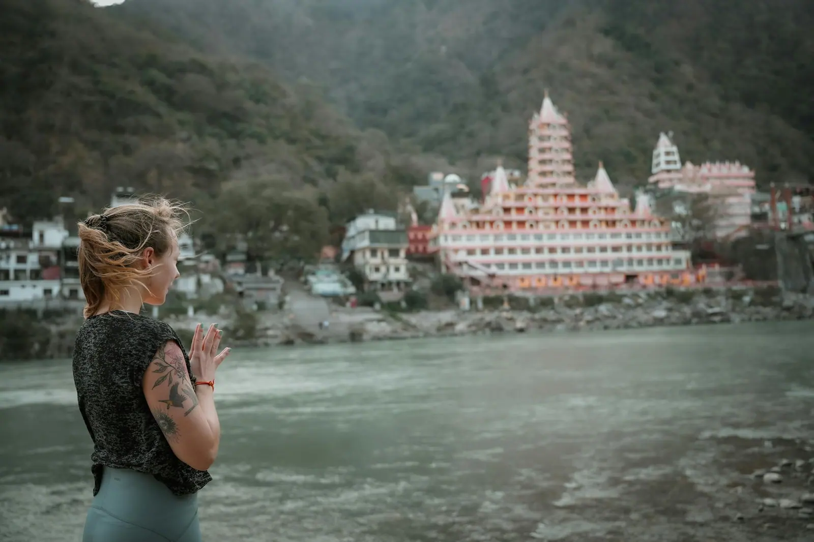 A woman practices yoga by the ganges river in rishikesh with a temple in the background