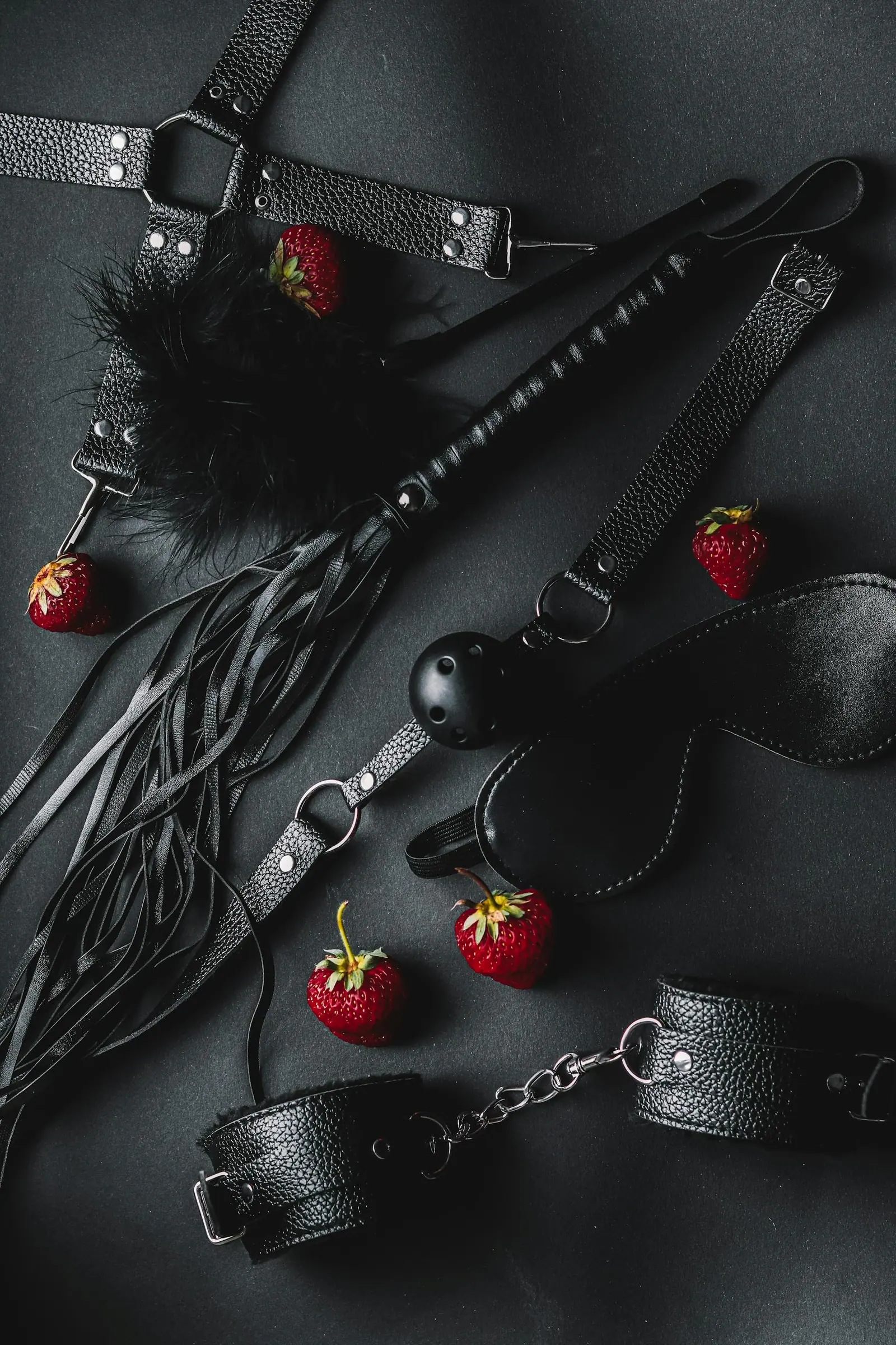A stylish flat lay of leather bdsm accessories with fresh strawberries on a dark background